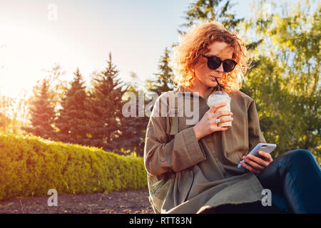Hipster young woman with curly red hair drinking coffee and using smartphone in summer park. Stylish girl chilling outdoors at sunset Stock Photo
