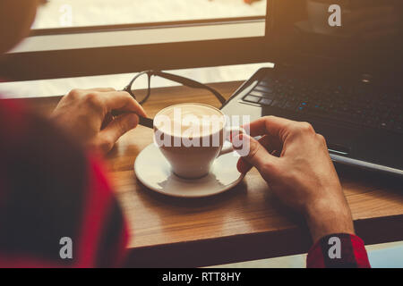 Young freelancer enjoys coffee in cafe using a laptop. Coffee break. Self-employed worker drinks coffee by window Stock Photo