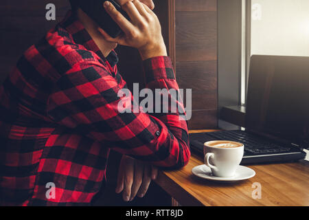 Young businessman enjoys coffee in the cafe talking on the phone and using a laptop in front of the window Stock Photo