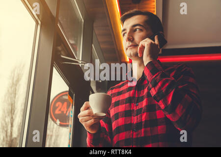Young man enjoys coffee in cafe talking on the phone by the window. Morning coffee before a working day Stock Photo