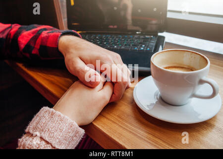 Young couple holds hands and enjoys coffee in the cafe while using a laptop in front of the window Stock Photo