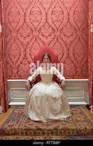 Queen Victoria sits on a throne at Kensington palace- the room is red and she wears a white dress Stock Photo