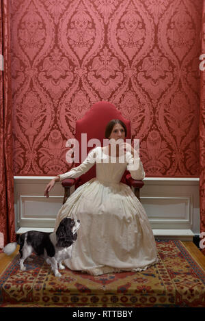 Queen Victoria sits on a throne at Kensington palace- the room is red and she wears a white dress. Her King Charles Spaniel called dash at her feet Stock Photo