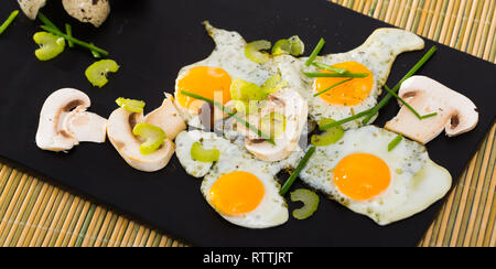 Broken fried quail eggs served on black board with mushrooms, sliced celery and onion Stock Photo