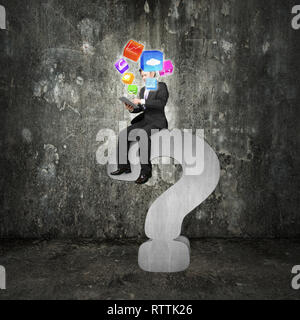 Businessman using tablet sitting on gray question mark with app icons and concrete room background Stock Photo