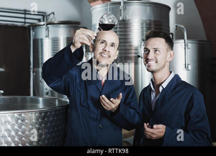 two cheerful winery male workers in uniform holding wine sample  in glass on factory Stock Photo