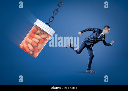 A side view of a businessman running from a huge full pills jar swinging on a chain on a blue background. Stock Photo