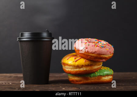 Three colorful donuts and coffee in a black paper Cup on a wooden table, on a black background. Bakery menu design concept. Hanukkah background. Stock Photo