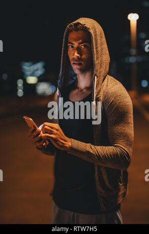 Fit young man in hooded shirt standing outdoors with mobile phone. fitness man taking break after workout outdoors at night. Stock Photo