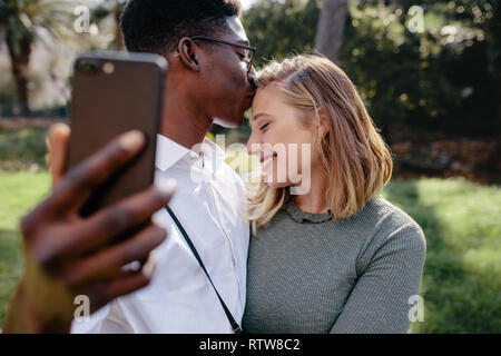 Loving couple taking selfie with smart phone outdoors. Man kissing on forehead of his girlfriend while taking self portrait.