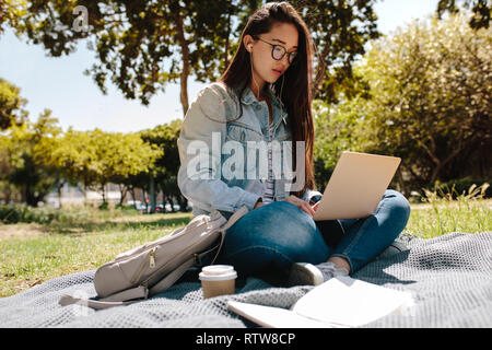 Close up of a female college student sitting in a lawn using her laptop computer on a sunny day. Asian college girl working on laptop sitting in a par Stock Photo