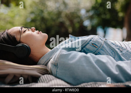 Side view of a woman relaxing outdoors listening to music wearing headphones. Close up of a young woman sleeping outdoors in a park enjoying the mild  Stock Photo