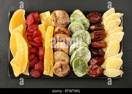 Dried fruit pieces of mango, papaya, kiwi; dried strawberries, figs, dates; pieces of candied ginger root, lying on a black ceramic plate on a black s Stock Photo