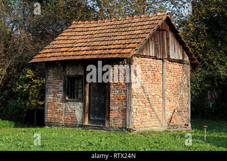 Backyard old red brick storage structure with dilapidated wooden doors and window surrounded with green grass and dense trees in background Stock Photo