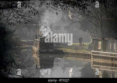 A misty morning on the Trent and Mersey canal at Great Haywood in Staffordshire. The scene shows dog walker and a boat with smoke coming from chimney Stock Photo