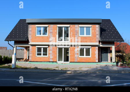 Family house under construction made of red building blocks with new doors and windows and unusual roof surrounded with gravel and paved road in front Stock Photo