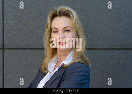 Confident smart businesswoman with folded arms standing against a grey wall smiling at the camera Stock Photo