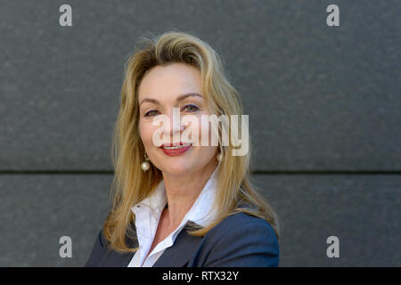 Attractive businesswoman with a friendly smile posing side on turning to face at the camera against a grey stone wall in a head and shoulders portrait Stock Photo