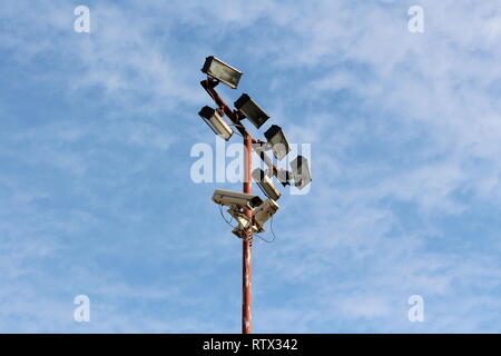 High metal pole with multiple light reflectors and security camera connected with electrical wires on cloudy blue sky background Stock Photo