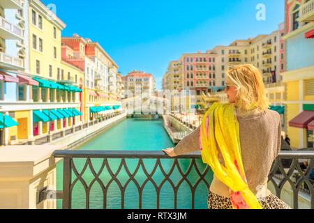Blonde woman on balcony looking canals of Venice, a Venetian style waterfront village in sunny day. Caucasian tourist enjoys Qanat Quartier in the Stock Photo