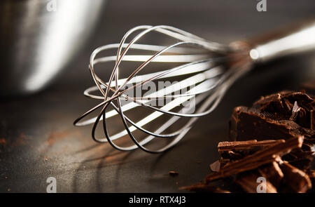 Chopped chocolate for baking alongside an old fashioned metal whisk on a wooden kitchen table in close up Stock Photo