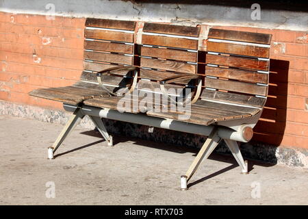 Vintage retro wooden public bench with three seats and two hand rests in middle mounted with strong metal frame on concrete in front of red brick wall Stock Photo