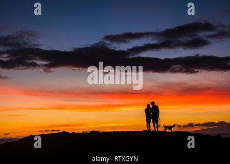 Romantic couple wioth dog enjoying and looking the sunset with red and orange coloured clouds in the sky - wanderlust and travel concept for happy peo Stock Photo