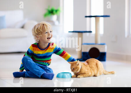 Child playing with cat at home. Kids and pets. Little boy feeding and petting cute ginger color cat. Cats tree and scratcher in living room interior.  Stock Photo