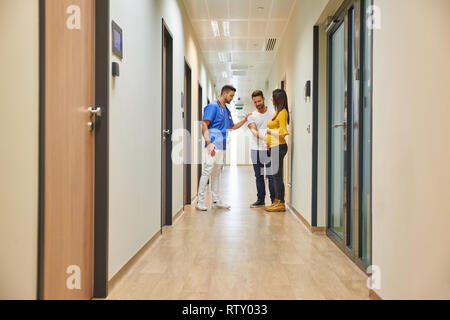 A handsome male doctor consulting with a young pregnant couple while standing in the hospitals hallway. Stock Photo