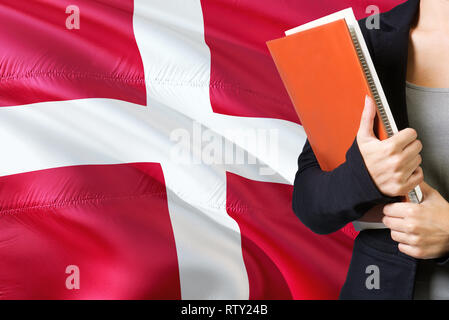 Learning Danish language concept. Young woman standing with the Denmark flag in the background. Teacher holding books, orange blank book cover. Stock Photo