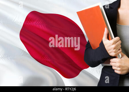 Learning Japanese language concept. Young woman standing with the Japan flag in the background. Teacher holding books, orange blank book cover. Stock Photo