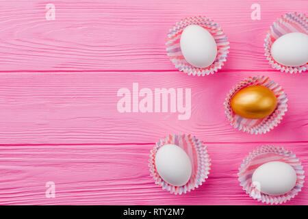 Eggs in paper wrapper, copy space. One gold egg and four white chicken eggs on pink wood with space for text. Easter greeting card. Stock Photo