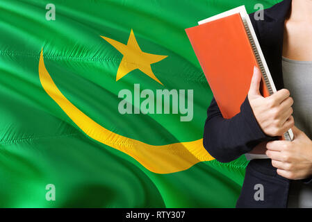 Learning Mauritanian language concept. Young woman standing with the Mauritania flag in the background. Teacher holding books, orange blank book cover Stock Photo