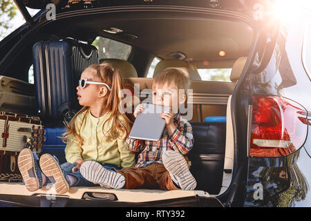 Two adorable caucasian kids sitting in a car trunk before going on vacations with their parents. Boy and girl looking forward for a road trip or trave Stock Photo