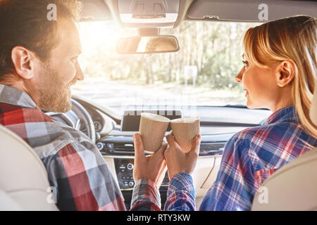 Smiling happy young couple, wearing checkered shirts, drinking coffee in paper cups, sitting together in a car Stock Photo