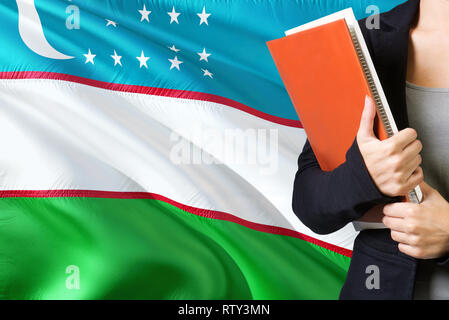 Learning Uzbek language concept. Young woman standing with the Uzbekistan flag in the background. Teacher holding books, orange blank book cover. Stock Photo