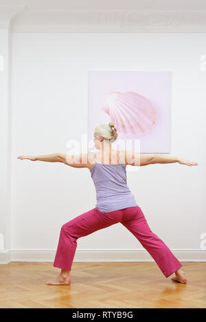 Senior woman with gray hair doing yoga exercises at home in front of a wall with a picture, asana warrior II Stock Photo