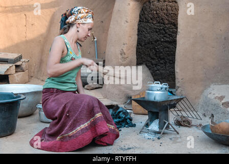 A caucasian woman cooking a meal in a traditional kitchen in rural Ghana, West Africa. Woman wears traditional Ghanaian clothes. Stock Photo