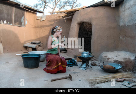 A caucasian woman cooking a meal in a traditional kitchen in rural Ghana, West Africa. Woman wears traditional Ghanaian clothes. Stock Photo