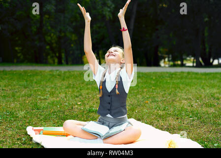 student in uniform smiles happily and raised  two hand above her head. schoolgirl is sitting on a blanket in a park with books and doing homework