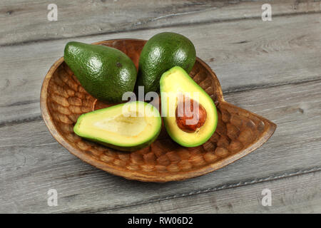 Three avocados, one of them cut to half, seed visible, in carved wooden bowl. Stock Photo