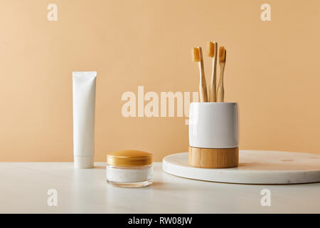 white holder with bamboo toothbrushes on marble round board, toothpaste in tube and cosmetic cream on table and beige background Stock Photo