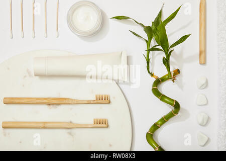 top view of marble round board with toothbrushes and toothpaste in tube, cosmetic cream, stones, ear sticks, bamboo stem on white background Stock Photo