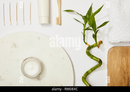 top view of marble round board with cosmetic cream and toothbrush, and toothpaste in tube, ear sticks, bamboo stem and towel on white background Stock Photo