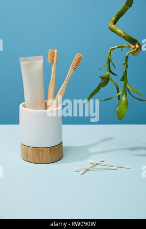 holder with bamboo toothbrushes, toothpaste in tube, ear sticks and bamboo stem on table and blue background Stock Photo