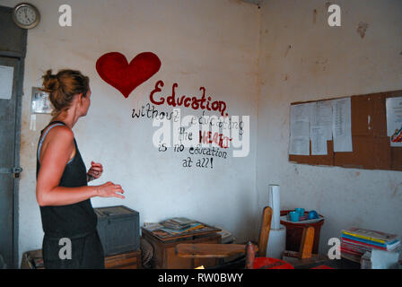 A caucasian woman standing on a local school office of a Ghanaian school. There is a beautiful heart and a message painted on the wall Stock Photo