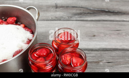 Three bottles with strawberries covered in syrup, large steel pot with sugar covered strawberry next to it. Home-made pickled fruit and marmalade prep Stock Photo