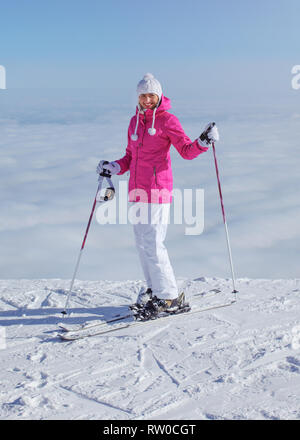Woman in pink jacket, ski poles and skis, standing at the edge of hill, only clouds below, looking back, smiling Stock Photo
