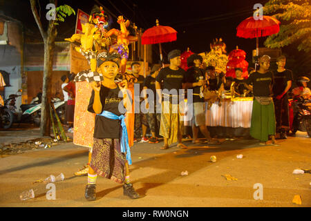 BALI, INDONESIA - MARCH 27, 2017: Boy in traditional costume holding Nyepi figures, traditional parade in evening Kuta street in background Stock Photo