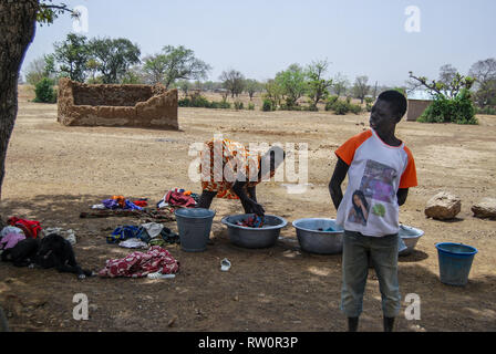 A ghanaian woman wearing traditional clothes is doing laundry while her son is standing and watching on the foreground. Stock Photo